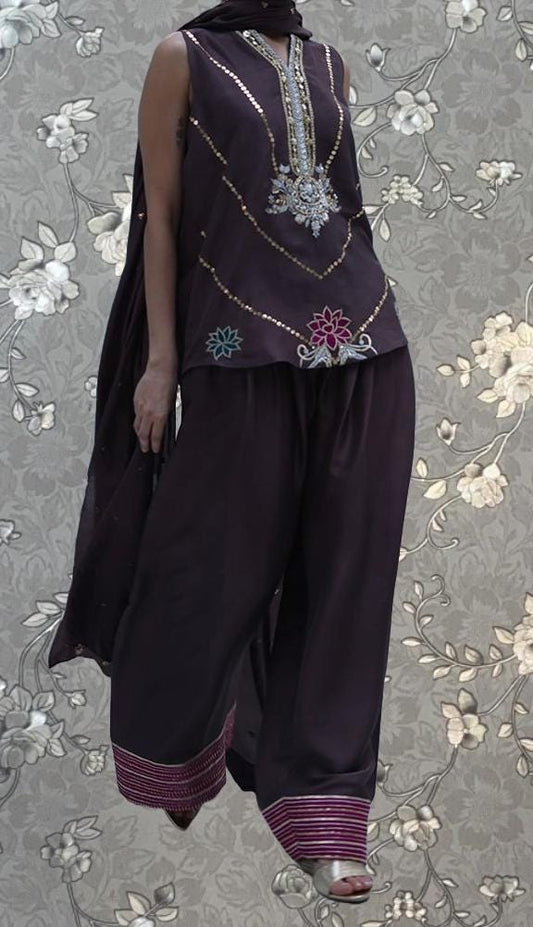 A person wearing a purple dress with intricate embroidery and floral patterns, standing against a floral background.