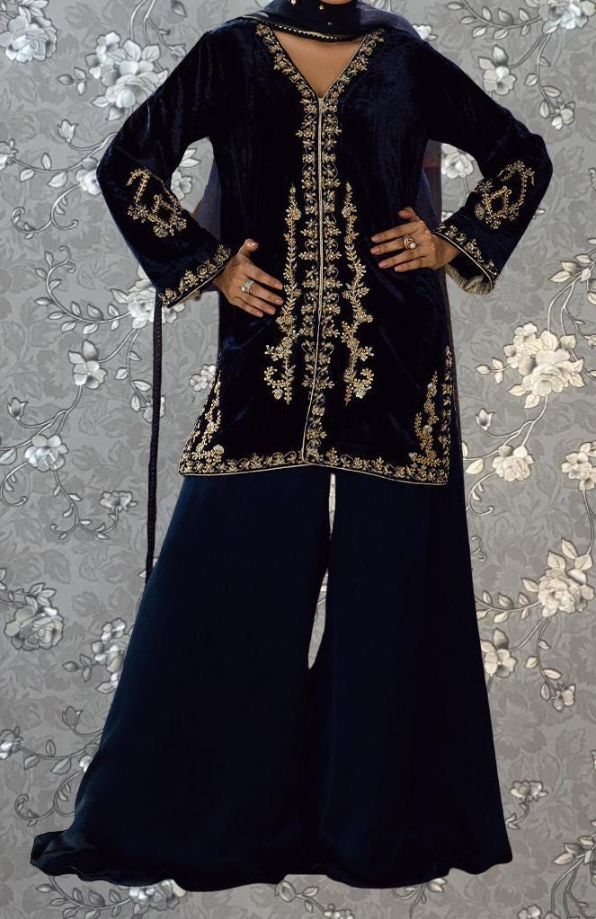 A person wearing a long, black velvet dress with gold embroidery and a matching black skirt, standing against a gray floral background.