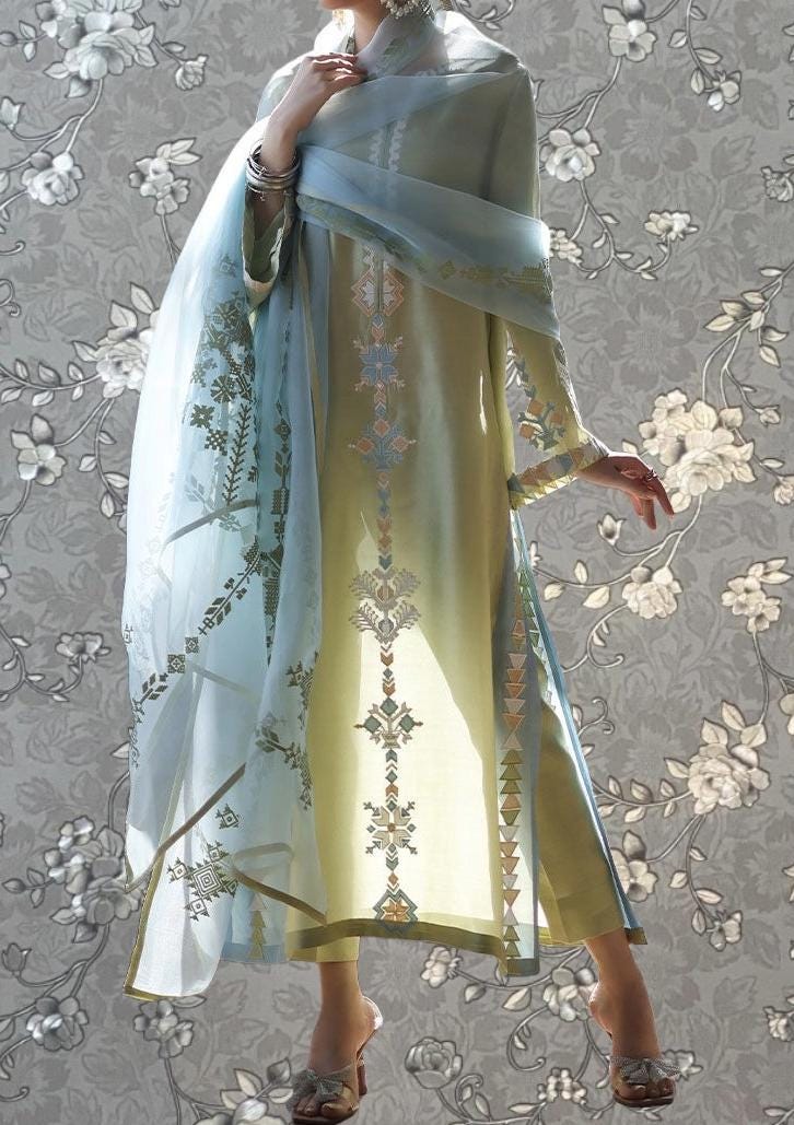 A person wearing a long, light-colored dress with intricate embroidered patterns, standing against a gray floral background.