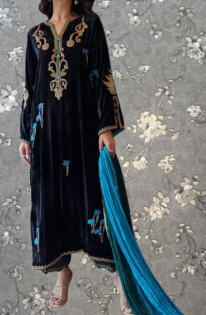 A woman wearing a long black dress with gold embroidery and a blue shawl is standing against a gray floral background.
