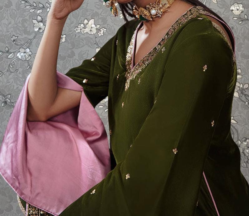 A woman wearing a green dress with gold embroidery and jewelry poses with her hand on her face.