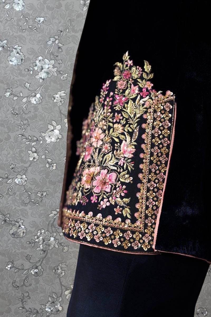 a close-up view of a black jacket with a floral pattern on the front. The jacket appears to be made of velvet and has a dark color. The floral pattern is intricate and detailed, featuring a variety of pink and white flowers. The background is a gray floral pattern, which complements the floral design on the jacket.