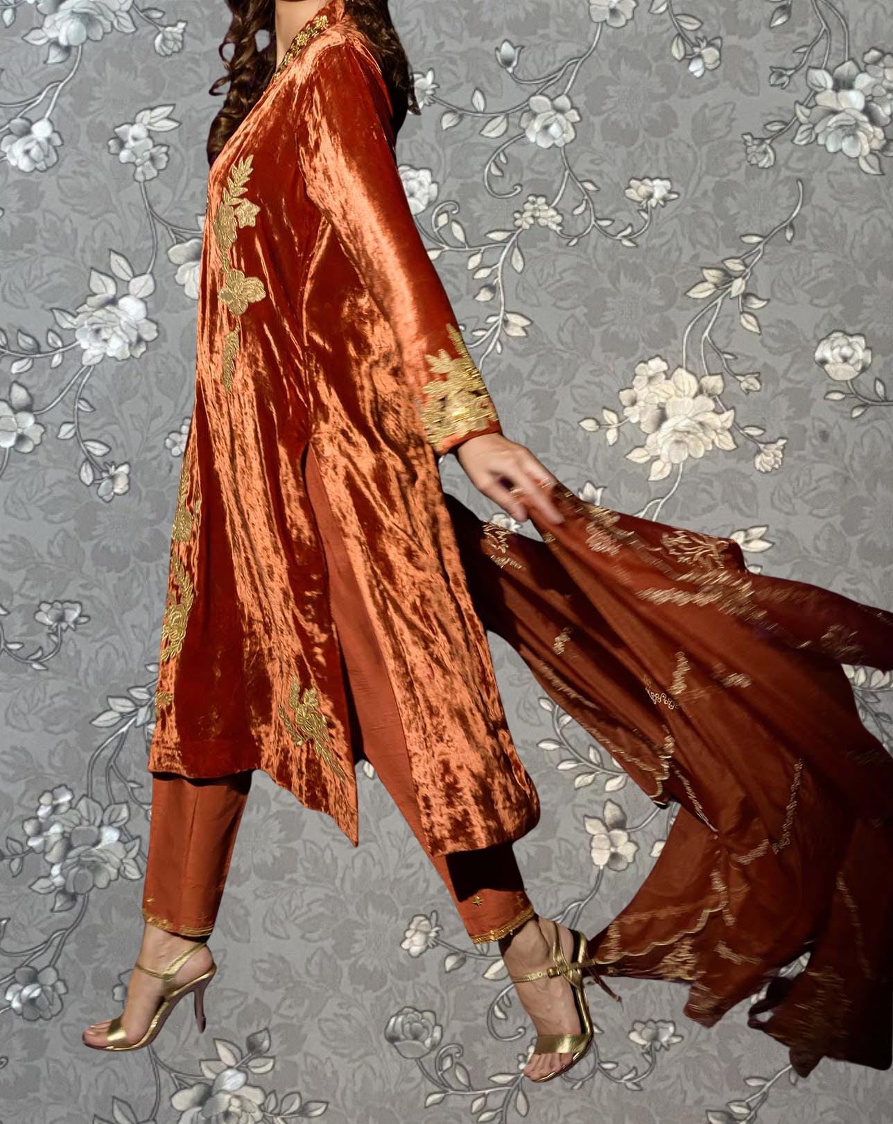 A woman wearing a long, red velvet dress with gold embroidery is shown against a gray floral background.