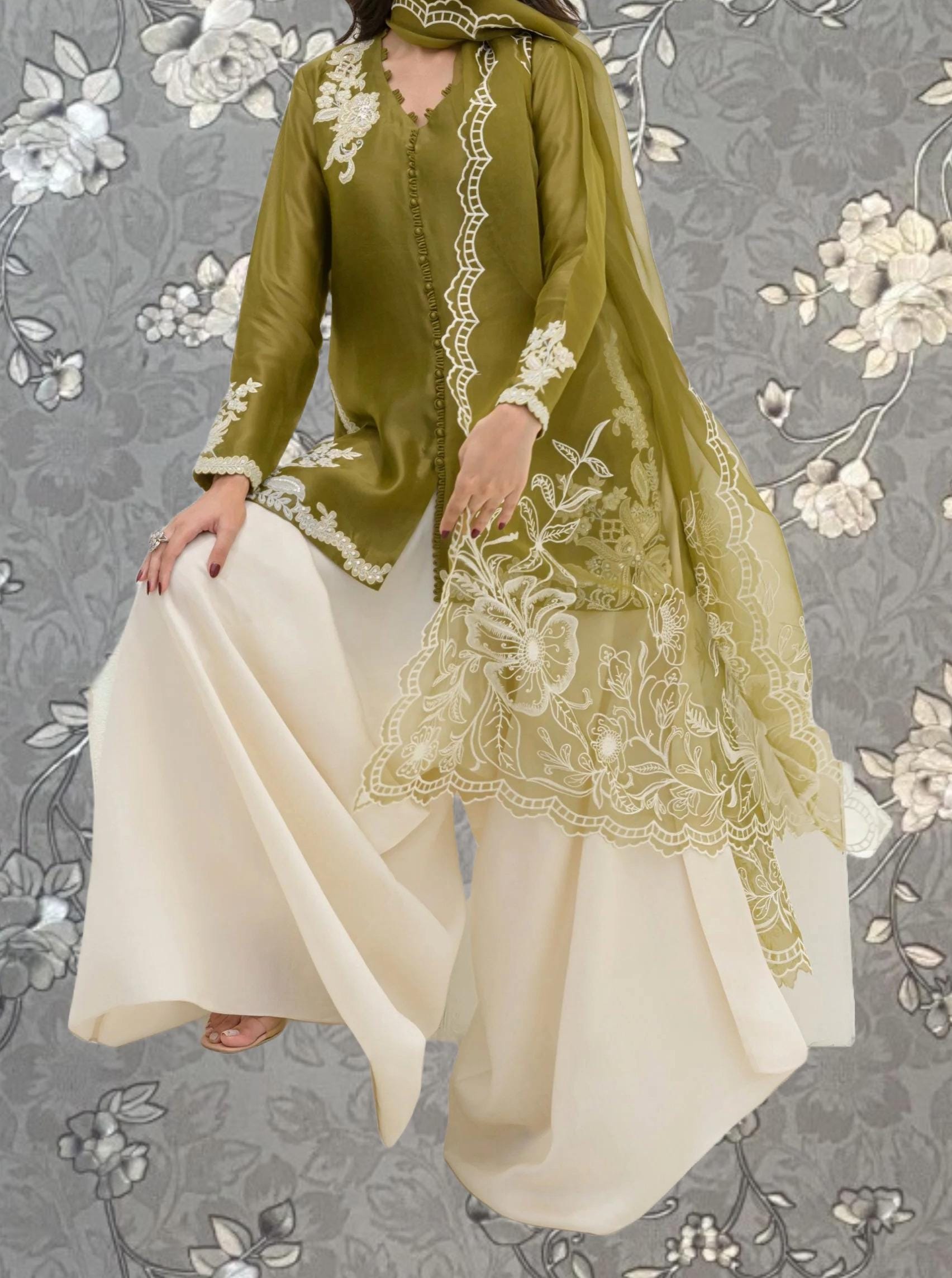 A woman wearing a green and white dress with intricate embroidery poses against a gray floral background.
