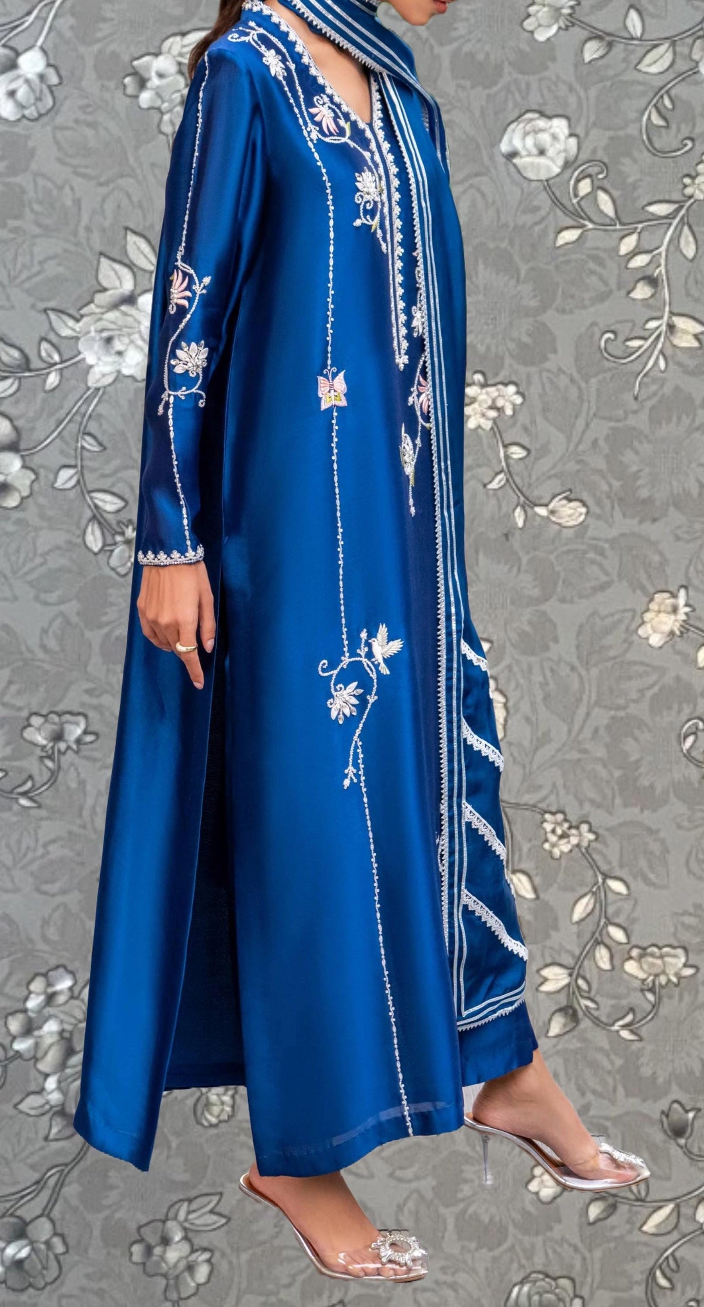 A woman wearing a long, blue dress with intricate embroidery and floral patterns, standing against a gray floral background.