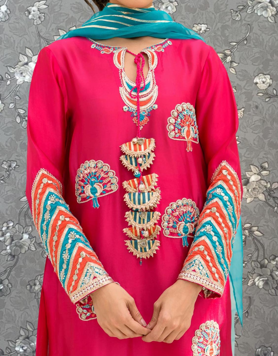A person wearing a bright pink dress with intricate embroidered designs and patterns, including peacock feathers, on the sleeves and collar.