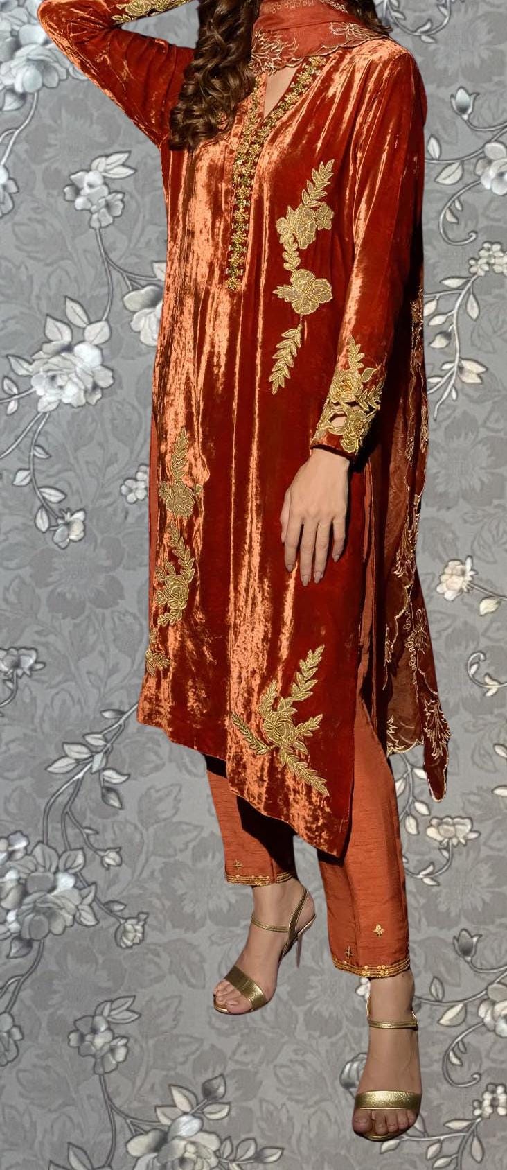 A woman wearing a vibrant red velvet dress with gold embroidery stands against a gray floral background.