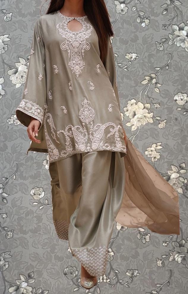 A woman wearing a grey and beige patterned dress with intricate embroidery, standing against a gray floral background.