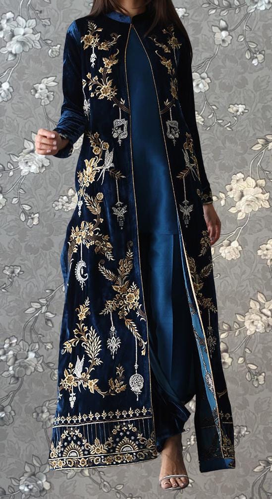 A woman wearing a long, blue velvet coat with gold embroidery stands against a gray floral background.