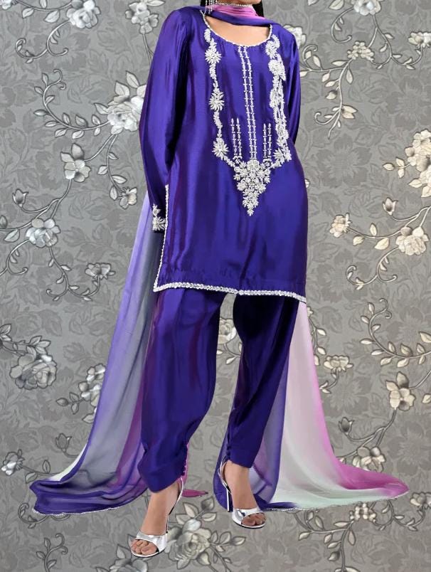 A woman wearing a purple and pink dress with intricate embroidery and a long train, standing against a gray floral background.