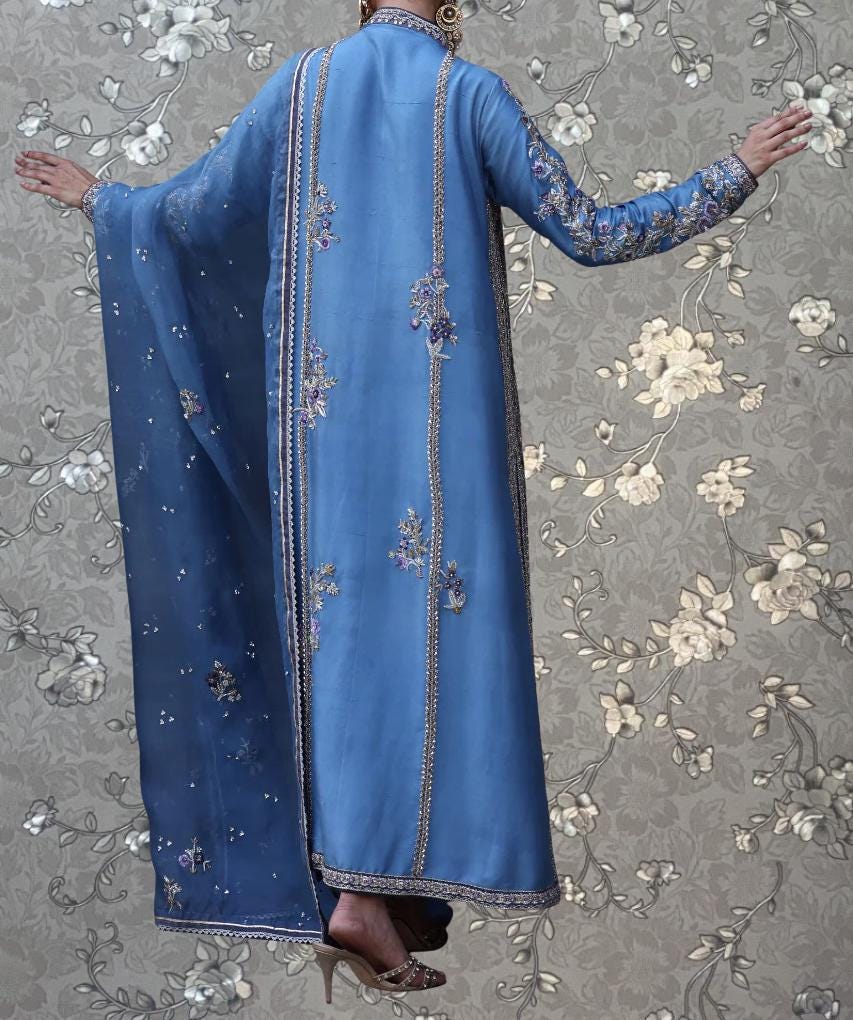 A person wearing a long, blue dress with intricate embroidery stands against a floral patterned background.