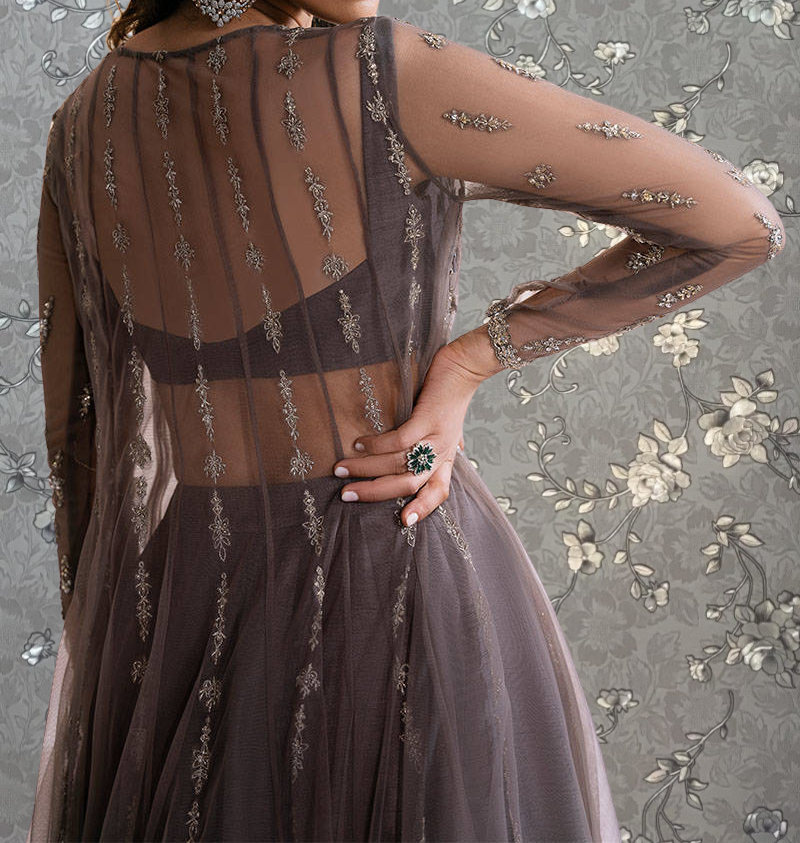 A woman wearing a long, sheer gray dress with intricate embroidery and a matching ring on her finger stands against a floral patterned background.