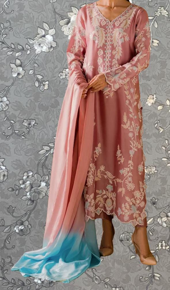 A woman wearing a pink and blue dress with intricate embroidery stands against a gray floral background.
