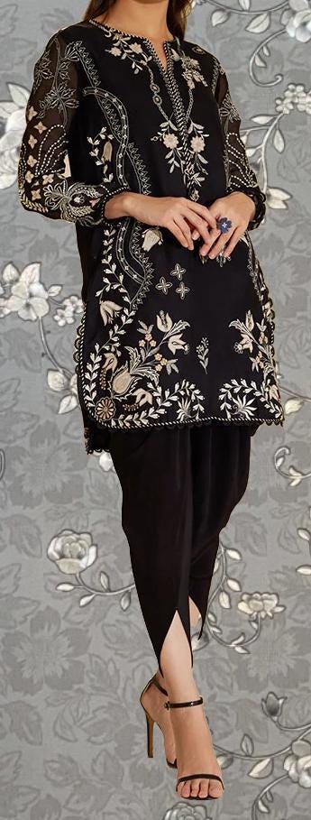 A woman wearing a black dress with intricate floral embroidery and high-heeled sandals stands against a gray floral background.