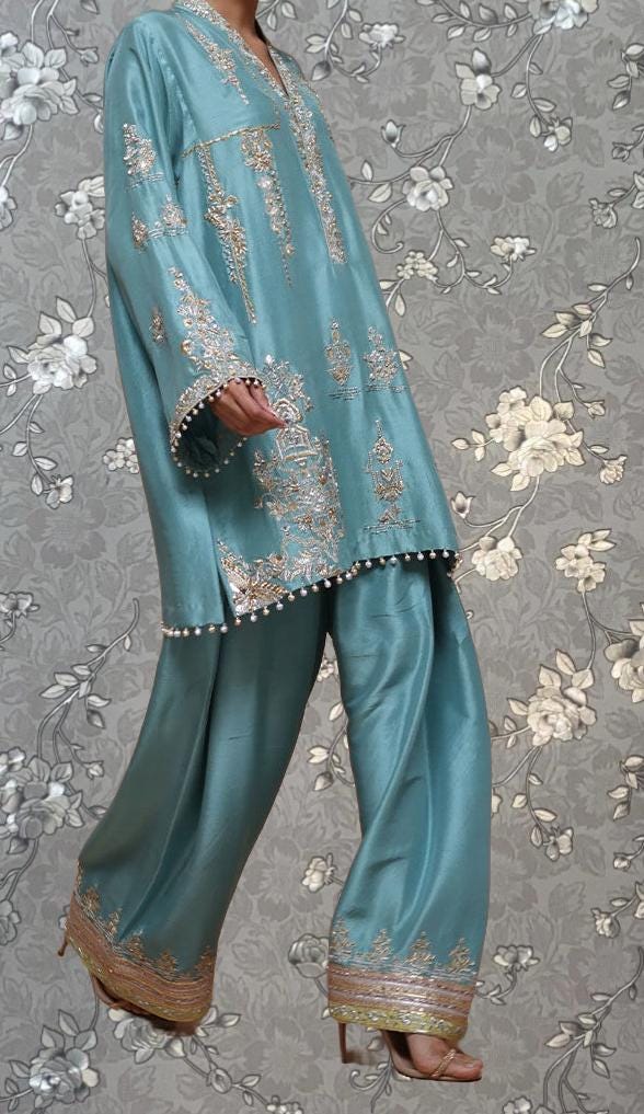 A person wearing a blue and gold patterned outfit, including a long-sleeved shirt and matching pants, stands against a gray floral background.