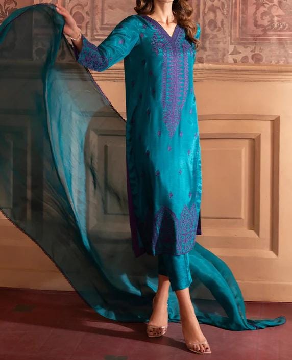 A woman wearing a teal and purple dress with intricate embroidery stands in front of a patterned wall.