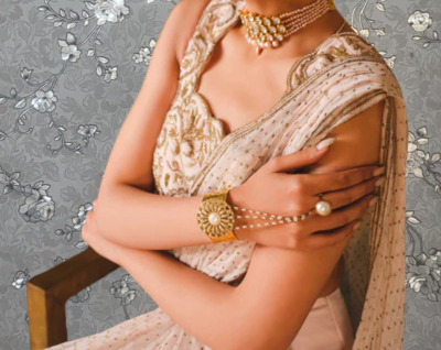 A woman wearing a white and gold dress with a large necklace and bracelet, posing with her arms crossed.