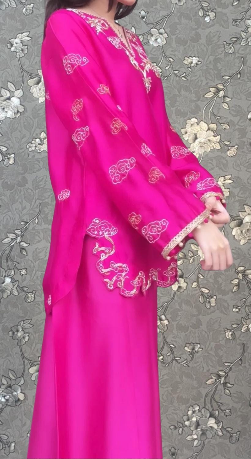 A woman wearing a long pink dress with intricate embroidery stands against a gray floral background.