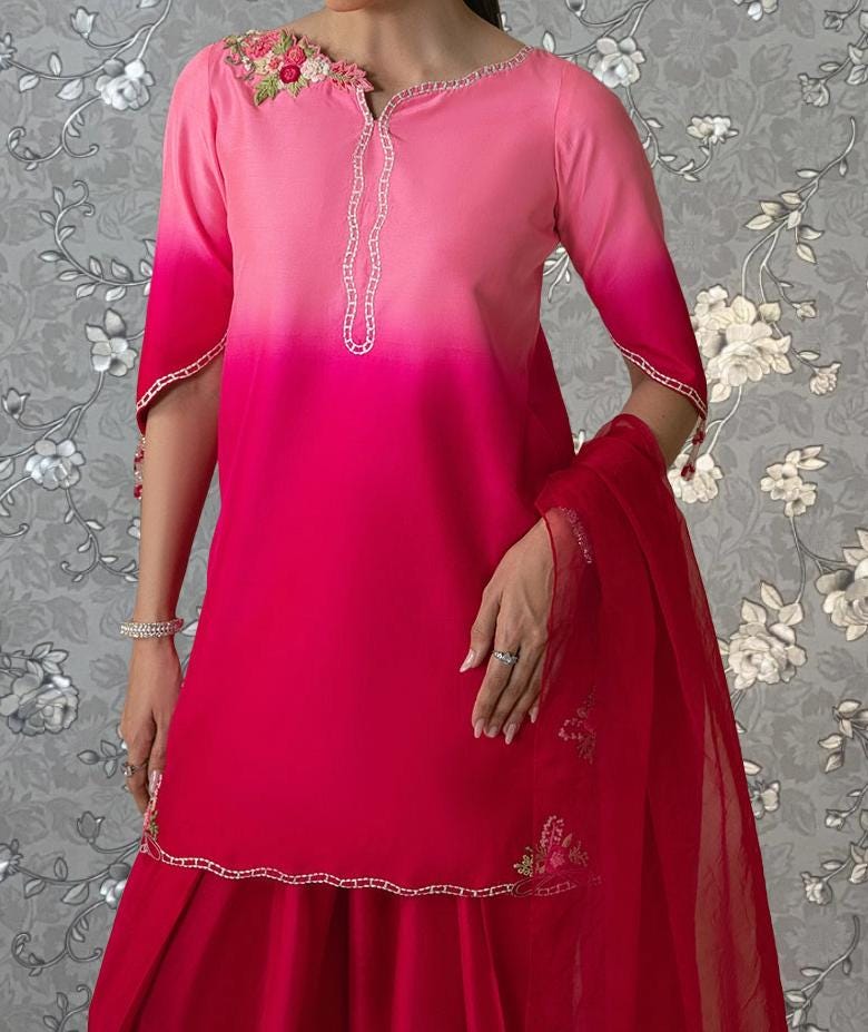 A woman wearing a pink and red dress with a matching red shawl, posing against a gray floral background.