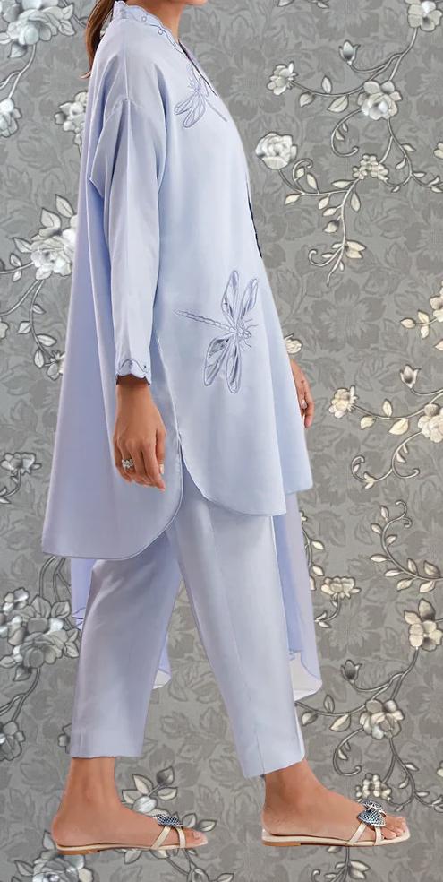 A person wearing a light blue outfit with a butterfly design on the left side, standing against a gray floral background.