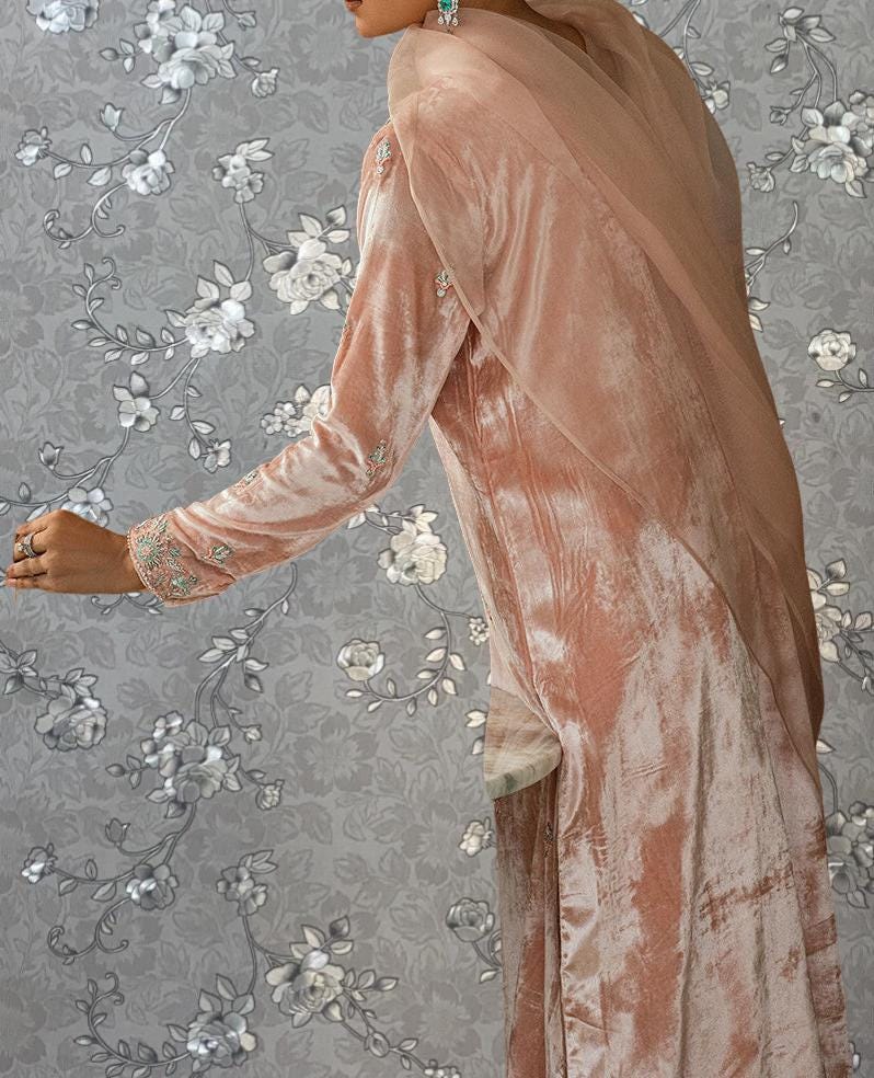 A woman wearing a long, peach-colored dress with a sheer overlay stands against a gray floral background.