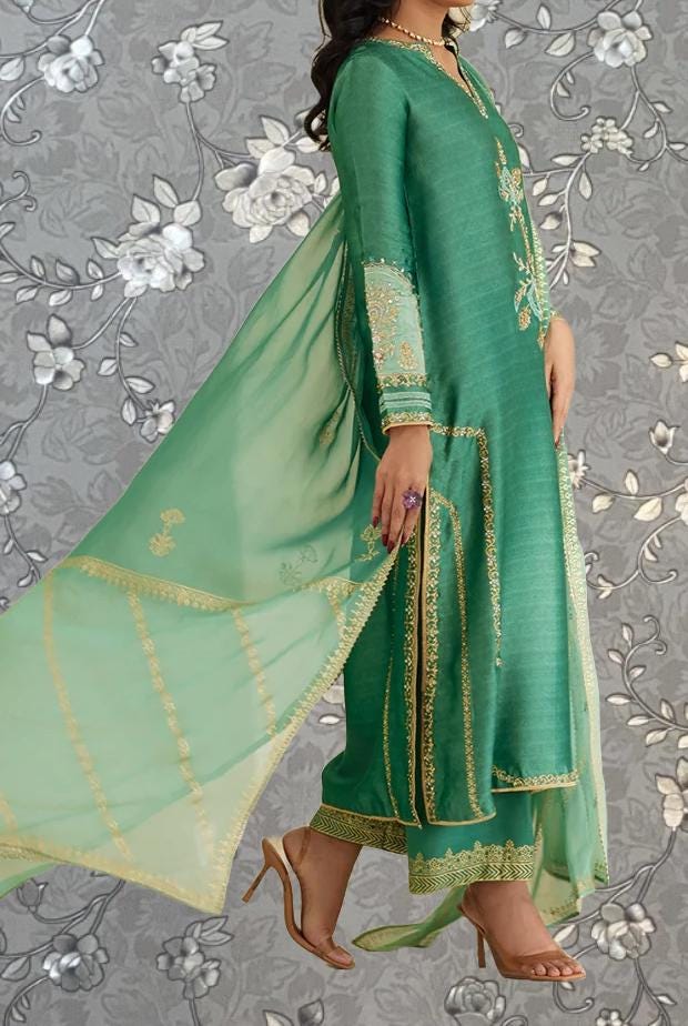 A woman wearing a green and gold dress with intricate embroidery, standing against a gray floral background.