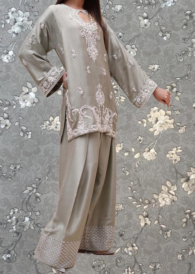 A woman wearing a gray dress with intricate embroidery stands against a gray floral background.