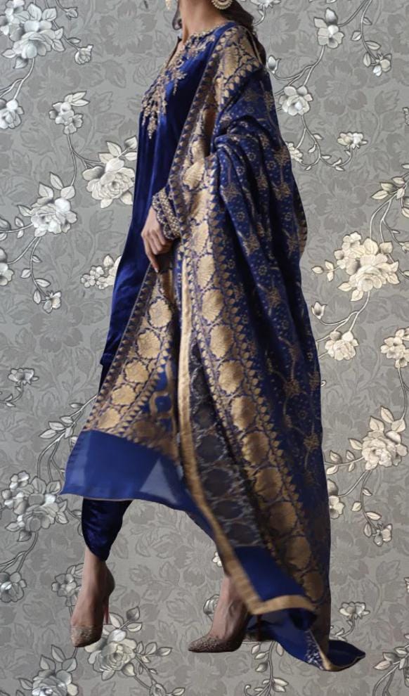 A woman wearing a blue and gold patterned shawl and matching blue and gold dress stands against a gray floral background.
