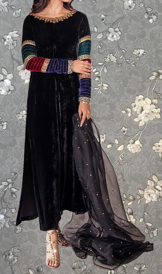 A woman wearing a long black velvet dress with colorful sleeves and a sheer black shawl draped over her shoulders, standing against a gray floral background.