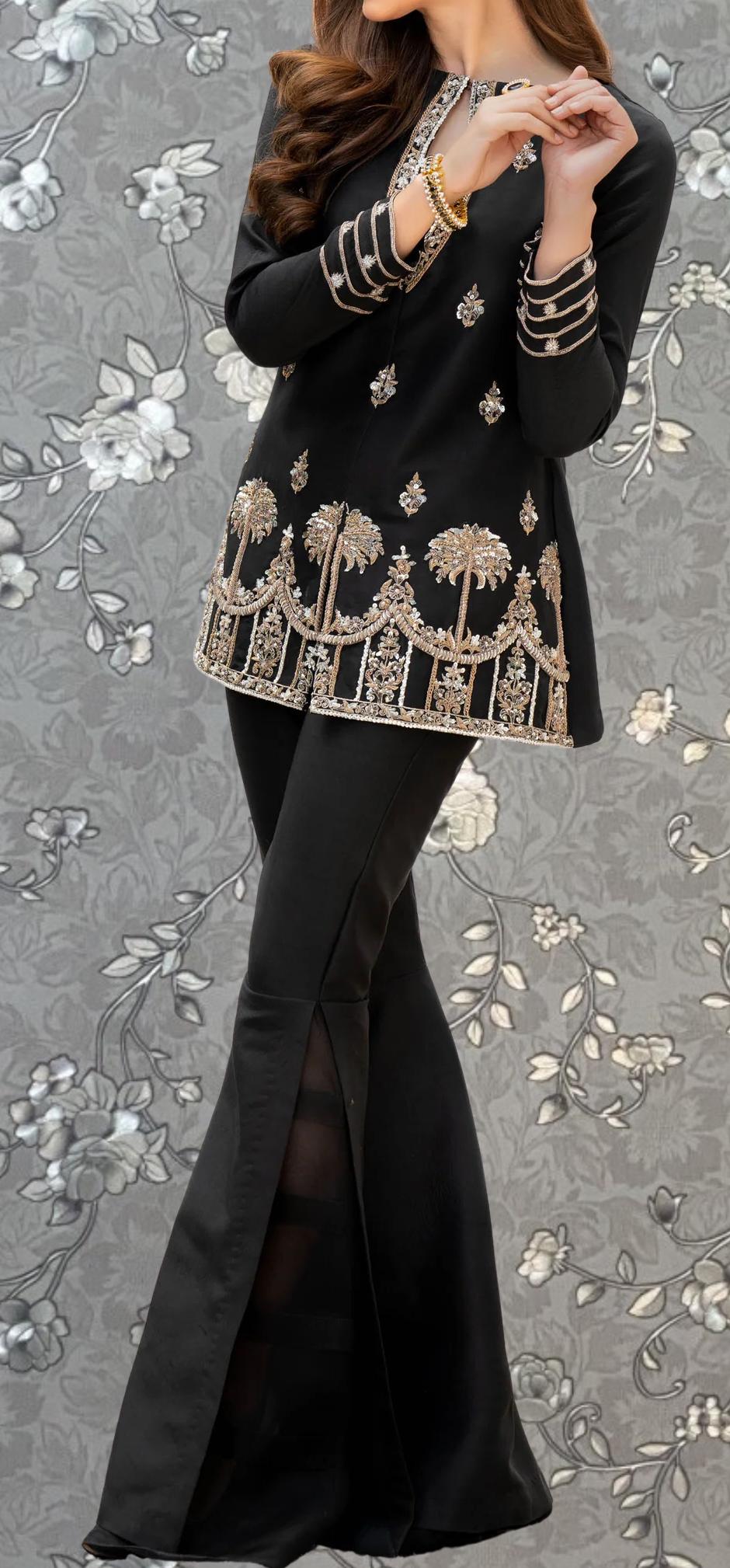 A woman wearing a black dress with gold embroidery and a floral pattern, standing against a gray floral background.