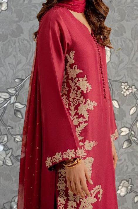 A woman wearing a red dress with gold embroidery stands against a gray floral background.