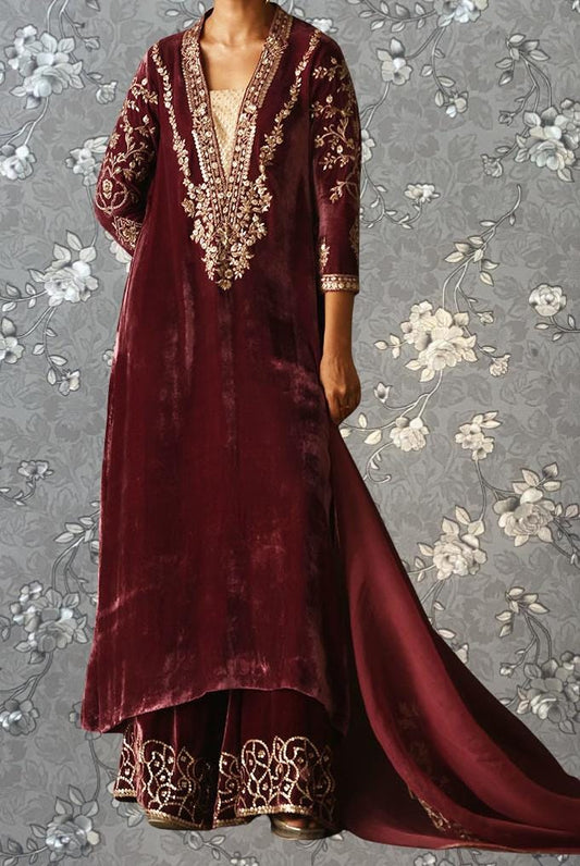 A person wearing a long, burgundy velvet dress with intricate gold embroidery and a matching skirt, standing against a gray floral background.