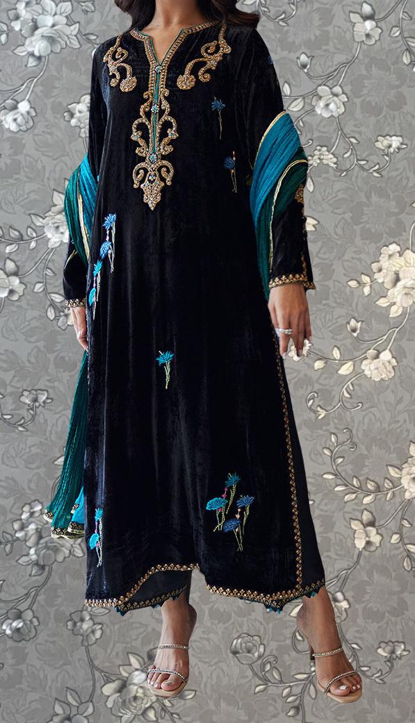 A woman wearing a long black dress with gold embroidery and blue accents, standing against a gray floral background.