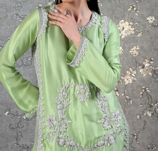 A woman wearing a green dress with intricate embroidery and lace details, posing against a floral patterned background.