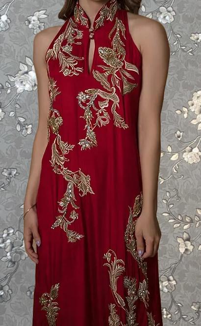 A woman wearing a red dress with intricate gold embroidery stands against a gray floral background.