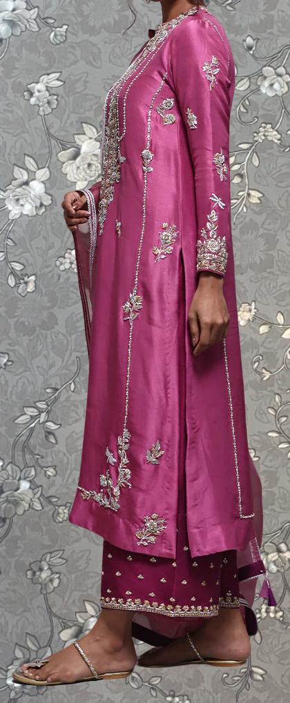 A woman wearing a long, pink dress with intricate embroidery stands against a gray floral background.