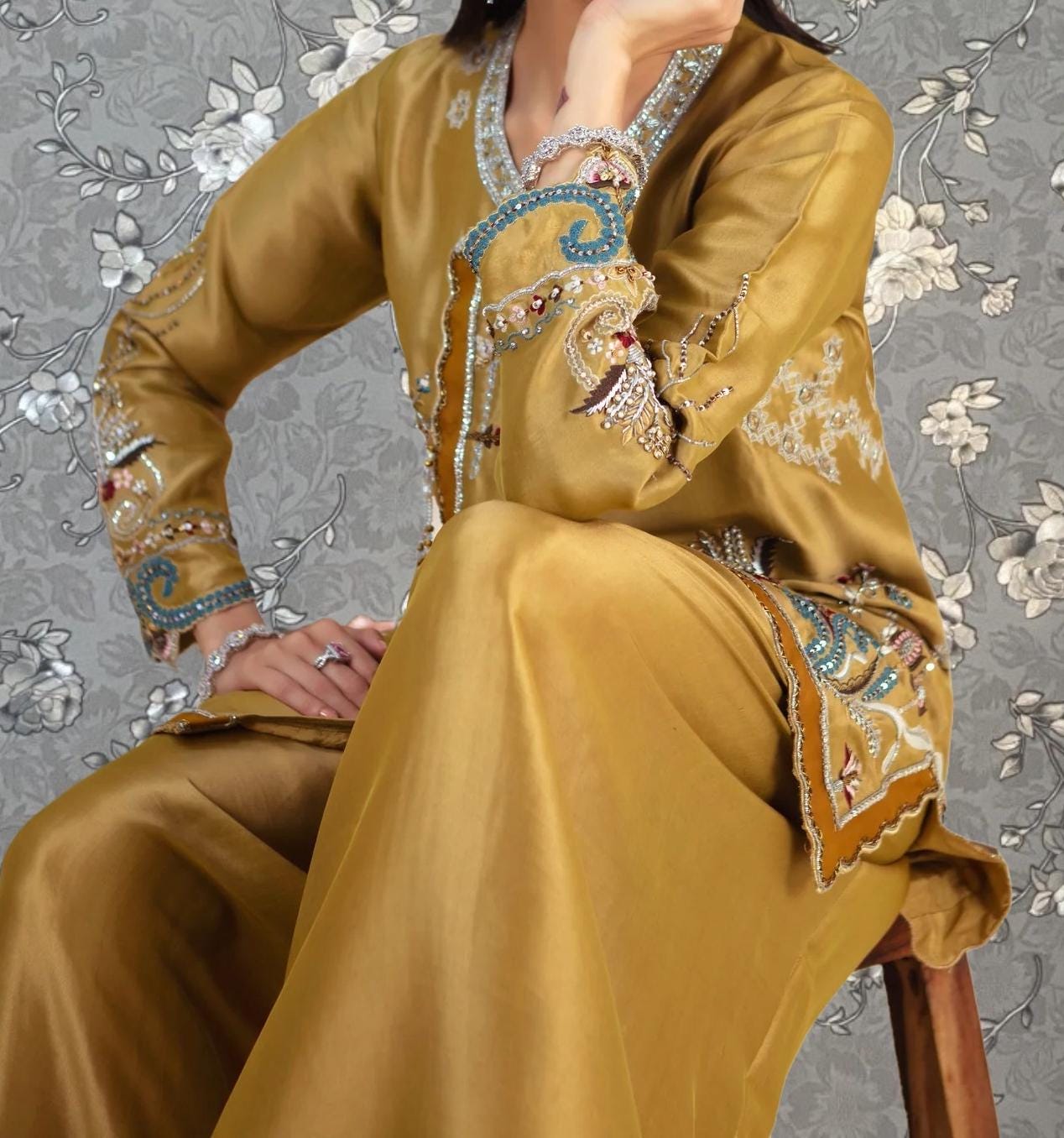 A woman wearing a gold and blue embroidered dress is sitting on a wooden stool against a gray floral background.