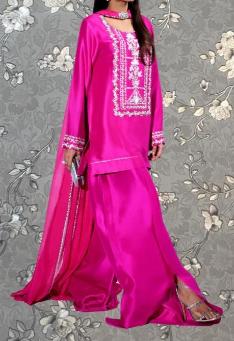 A woman wearing a long pink dress with intricate embroidery and a matching pink skirt, standing against a gray floral background.
