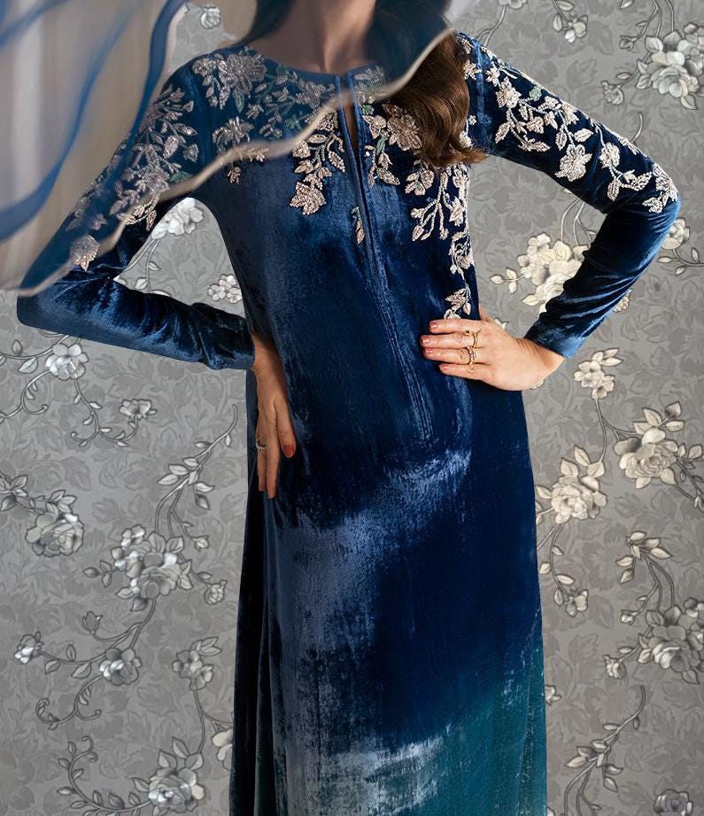 A woman wearing a long, blue velvet dress with white floral embroidery stands against a gray floral background.