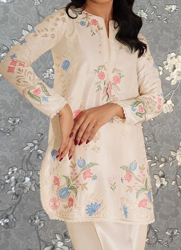 A woman wearing a white dress with floral embroidery poses against a gray floral background.