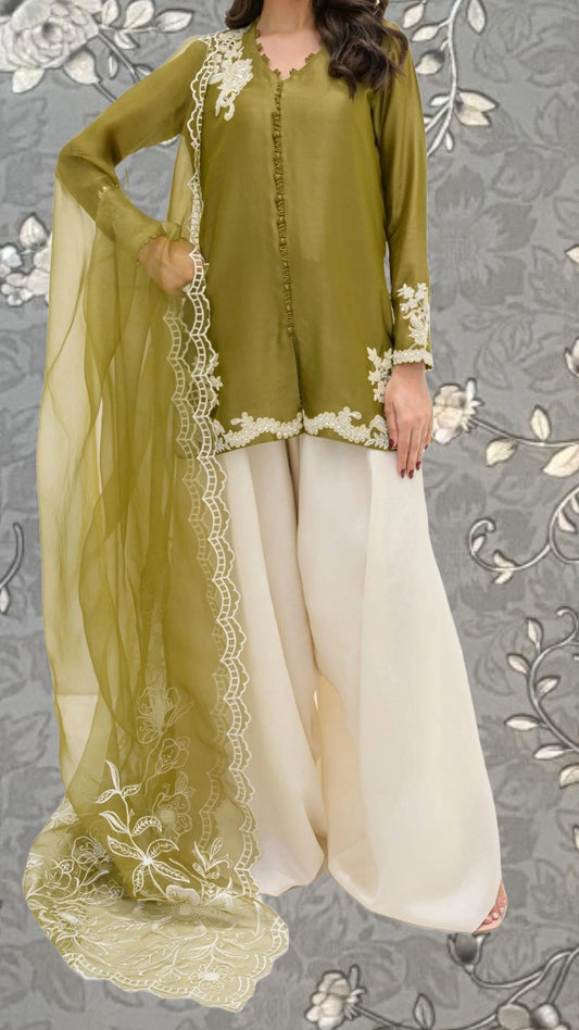 A woman wearing a green blouse with white embroidered details and a matching green shawl draped over her shoulders, standing against a gray floral background.