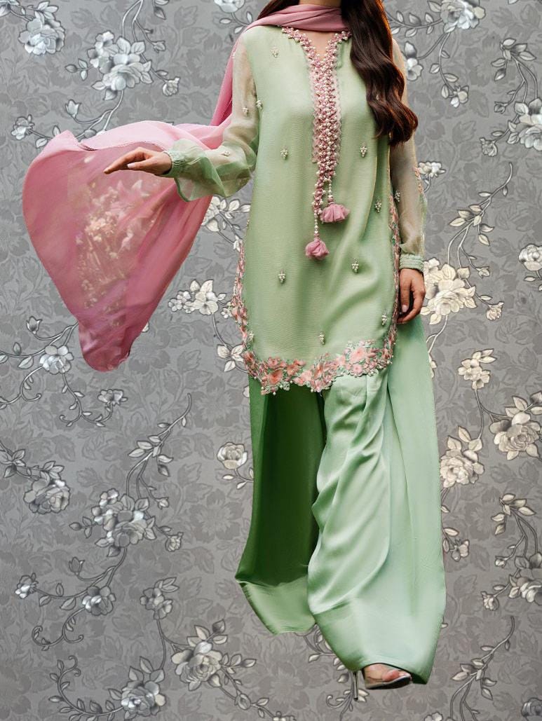 A woman wearing a green and pink dress with floral embroidery and a pink shawl is standing against a gray floral background.