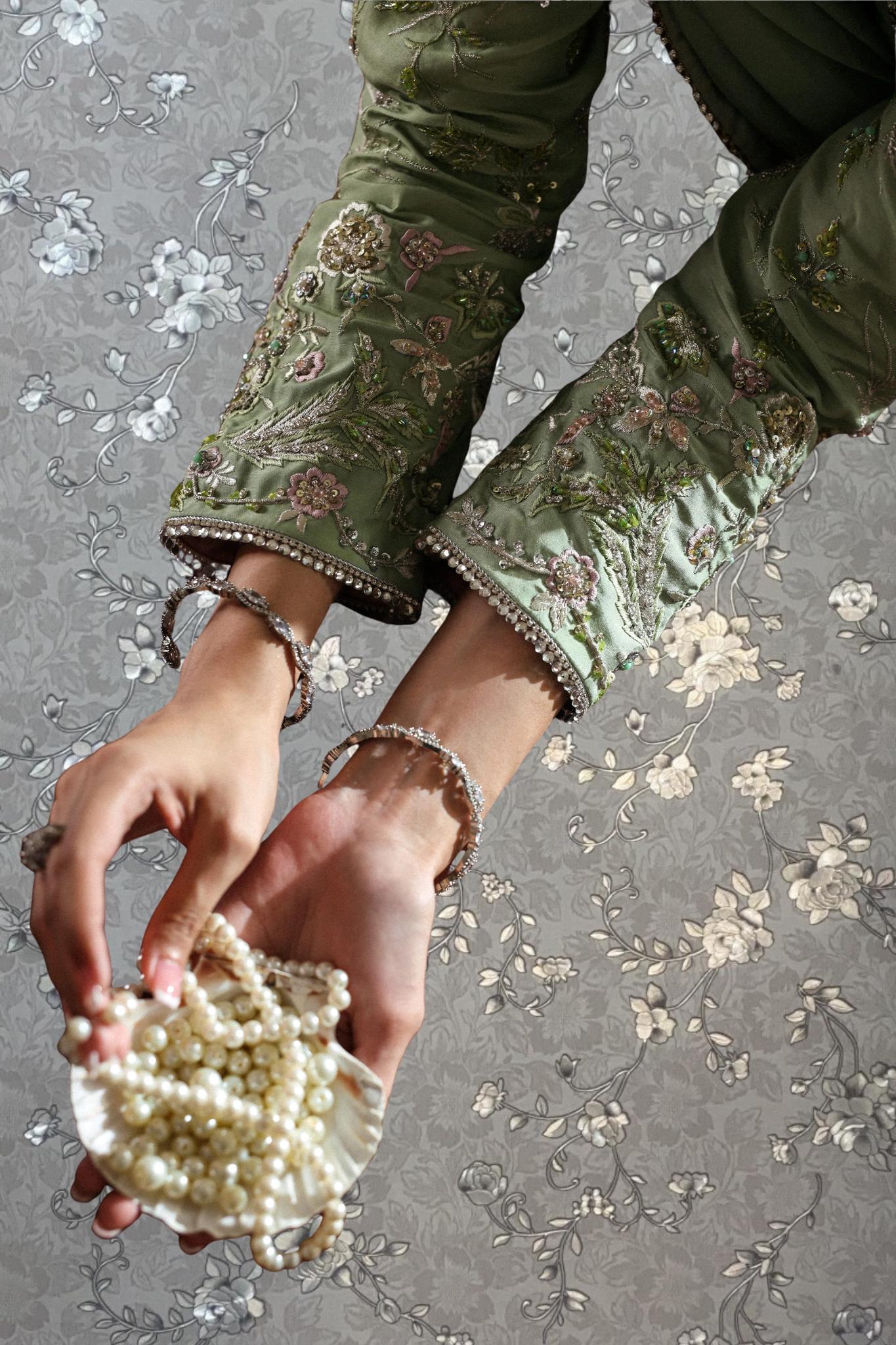 A person's hands holding a small white object, possibly a pearl, and wearing a green embroidered blouse with a floral pattern.