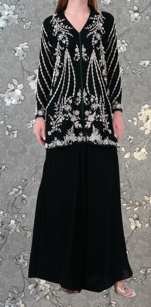 A woman wearing a black dress with white floral embroidery and a black jacket with white embroidery stands against a gray floral background.