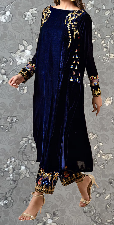 A woman wearing a long navy blue velvet dress with gold embroidery and floral patterns, paired with gold heels, stands against a gray floral background.