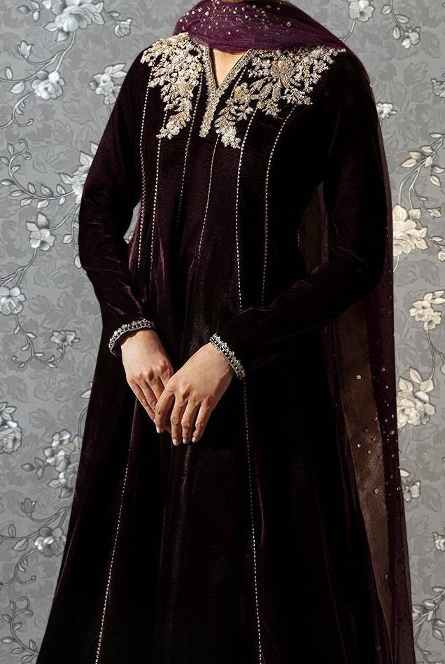 A person wearing a long, dark purple velvet dress with intricate embroidery and jewelry, standing against a floral background.