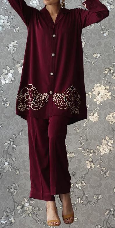 A person wearing a burgundy outfit with floral embroidery on the sleeves and pants, standing against a gray floral background.