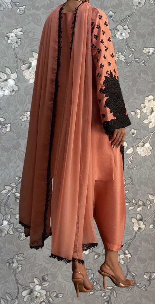 A person wearing a peach-colored dress with black floral accents, standing against a gray floral background.