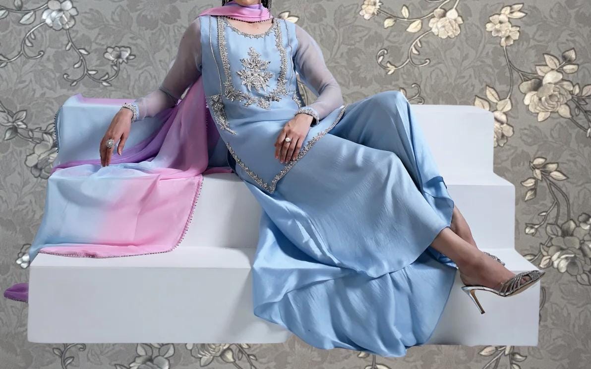 A woman wearing a blue dress with a pink and purple pattern is sitting on a white platform against a floral background.