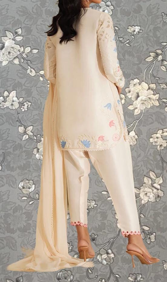 A woman wearing a cream-colored dress with floral embroidery stands against a gray floral background.